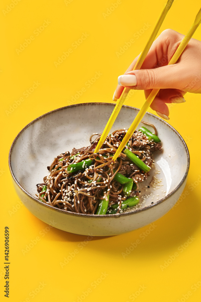 Woman using yellow chopsticks eating soba noodles with beef and green beans on vibrant yellow background