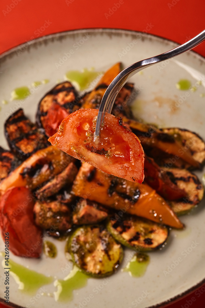 Fork lifting grilled tomato slice from plate of vegetables