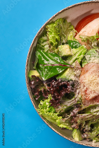 Fresh salad with chicken, avocado, tomatoes, and greens in bowl on blue background