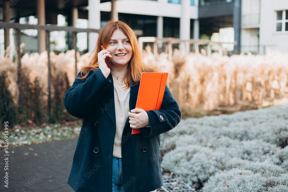 Young adult smiling professional business woman holding smartphone and folder with files while walking in urban city street.