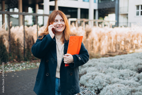 Obraz na płótnie Young adult smiling professional business woman holding smartphone and folder with files while walking in urban city street. w salonie