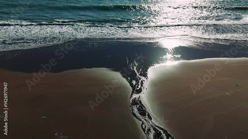 Sunlight gleaming on wet shoreline, stream merging with ocean waves, creating sparkling coastal landscape at golden hour