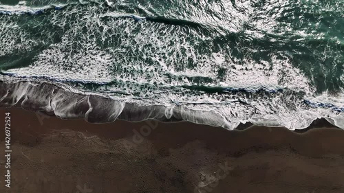 Aerial drone perspective revealing turquoise waves rhythmically breaking against dark sandy shoreline, generating hypnotic oceanic motion