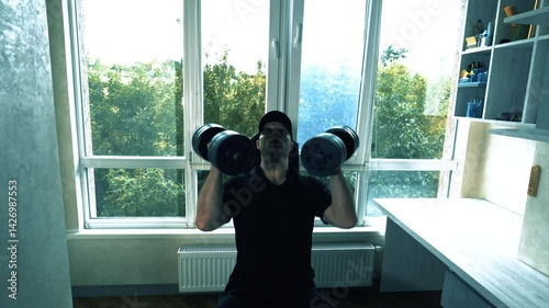 An athlete performs a shoulder exercise by lifting dumbbells with weight. Home workout. Young athlete in a workout apartment.