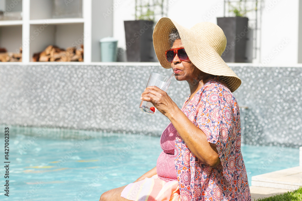 Senior woman sitting at poolside, wearing straw hat, red sunglasses, sipping from glass, copy space