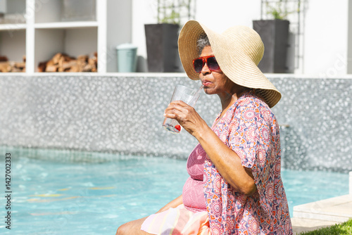 Obraz na płótnie Senior woman sitting at poolside, wearing straw hat, red sunglasses, sipping from glass, copy space w salonie