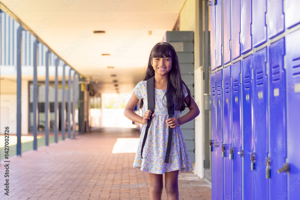 Multiracial child girl standing in school hallway, holding backpack straps with blue lockers