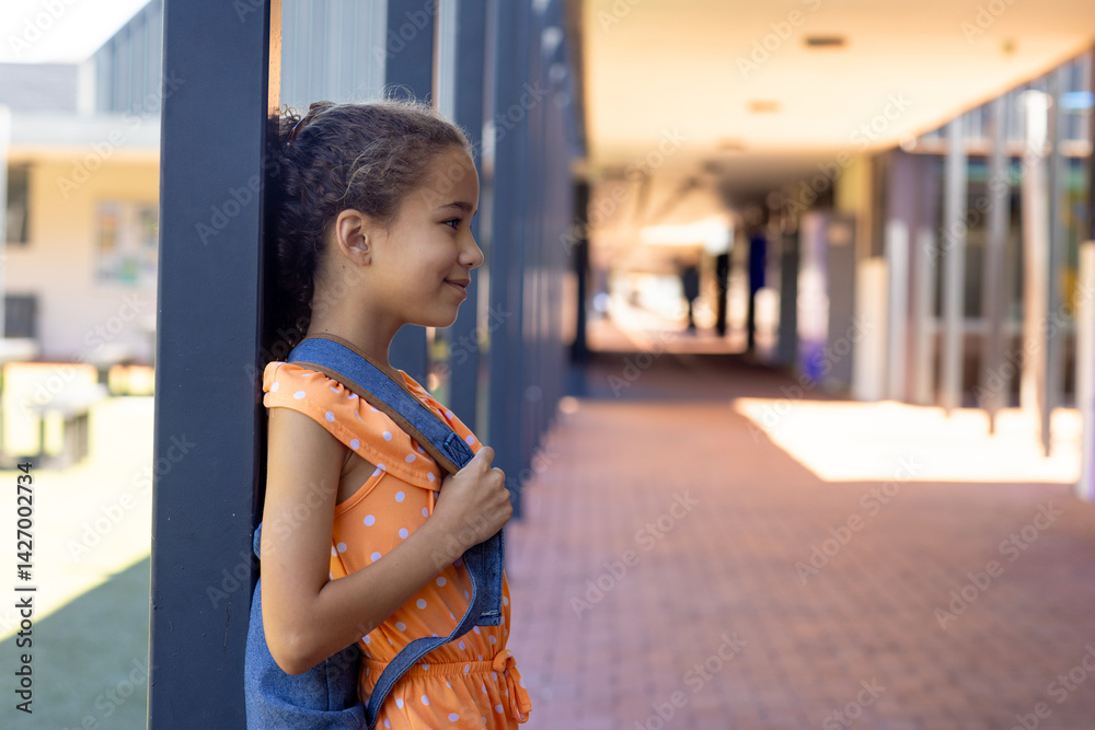 Girl child standing in profile at covered school walkway, holding denim backpack strap, copy space