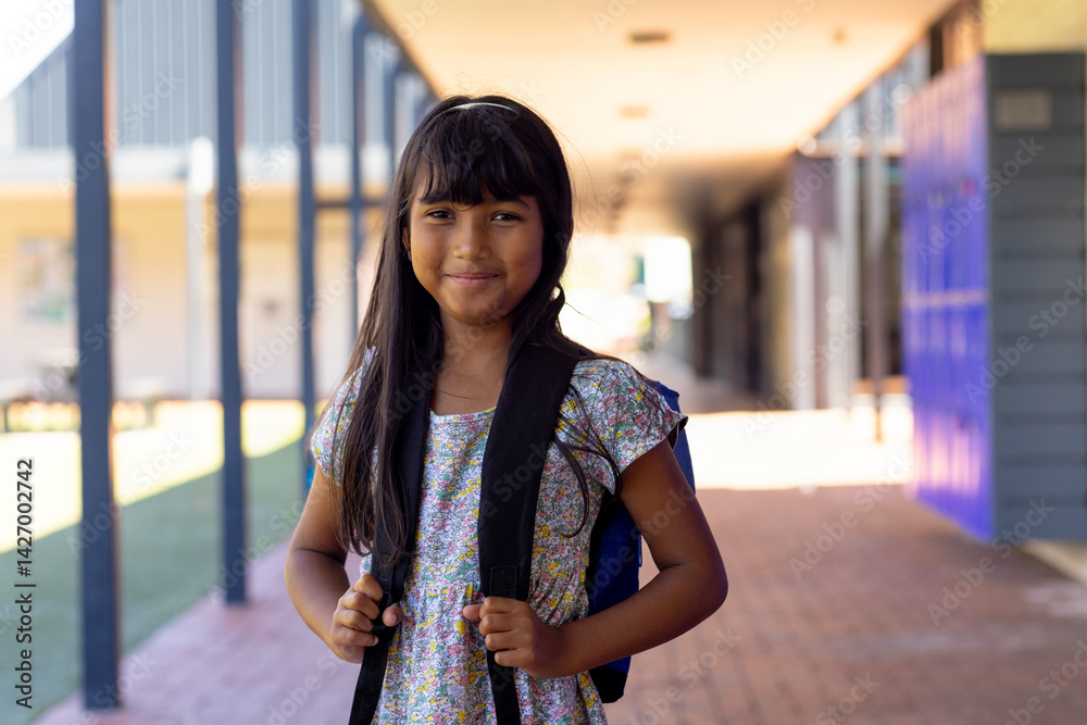8-year-old girl wearing floral dress standing in sunlit school corridor, gripping blue backpack