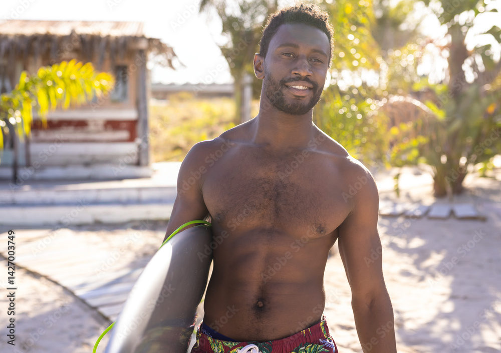 Young adult African American man standing on tropical beach holding surfboard, enjoying sunshine