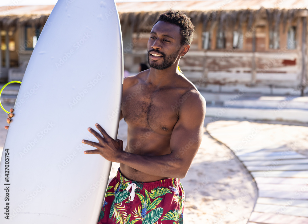 African American man on sandy beach near shack holding white surfboard, wearing red trunks