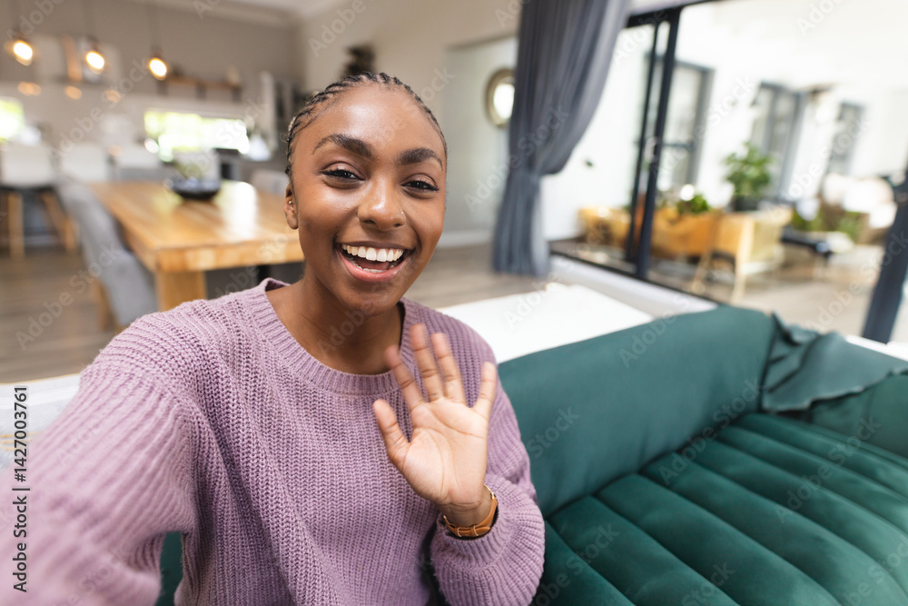 African American woman sitting on green sofa in open-plan living room, waving at camera, copy space