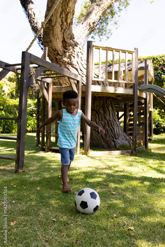 Young African American boy playing soccer in sunlit backyard, with soccer ball and wooden playset