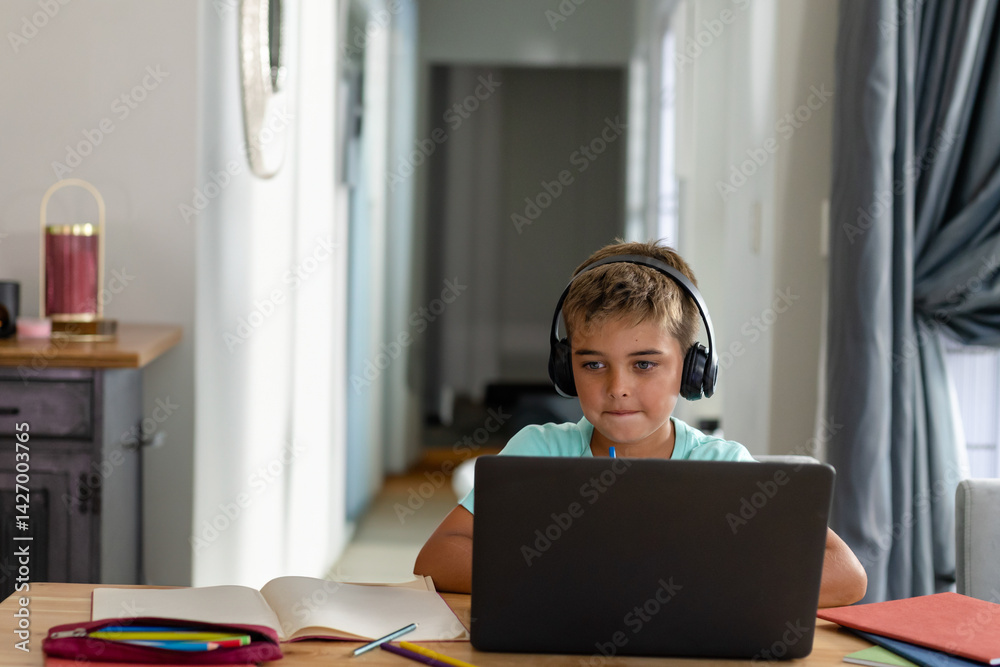 Boy sitting at wooden table in home study, using laptop with headphones and open notebook