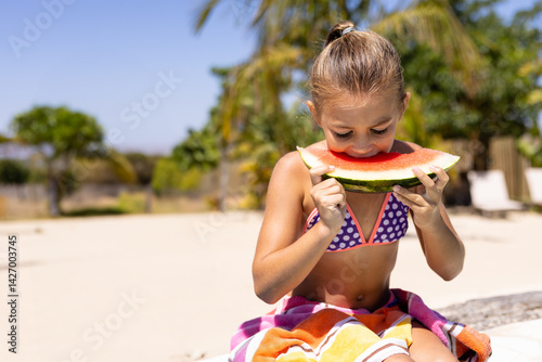 Child girl biting large watermelon slice while sitting on colorful towel on bench in backyard