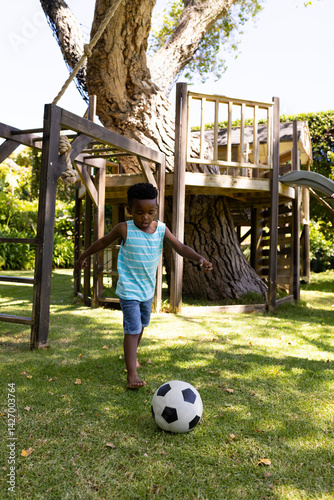 Obraz na płótnie Young African American boy playing soccer in sunlit backyard, with soccer ball and wooden playset w salonie