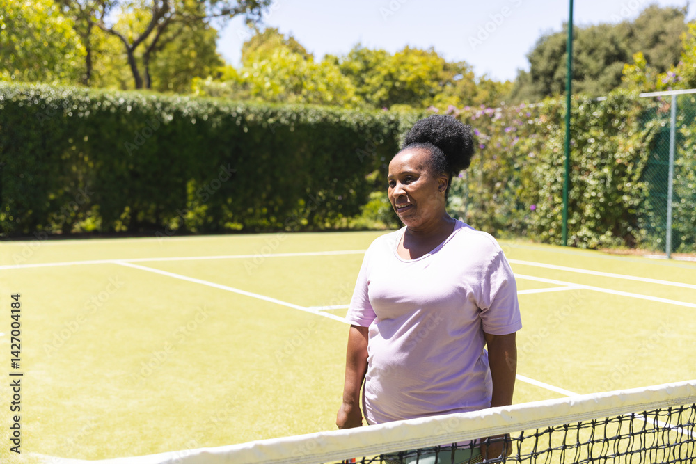 Senior African American woman standing and smiling on tennis court, with tennis net, court lines