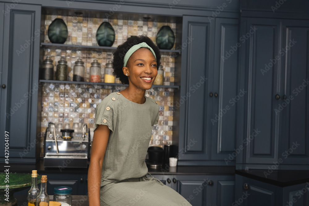 African American woman sitting on kitchen island, surrounded by oil bottles and mosaic backsplash