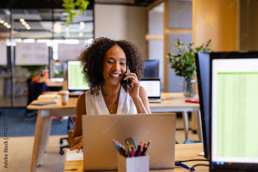 Woman working on silver laptop at modern open-plan office desk, smiling while holding smartphone