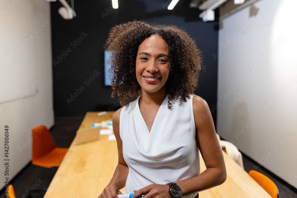 Woman leaning on table in office meeting room, holding whiteboard marker and smiling at camera