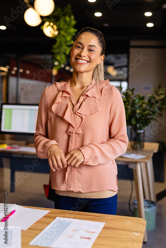 Woman in 30s standing behind wooden desk in modern office, smiling and reviewing charts