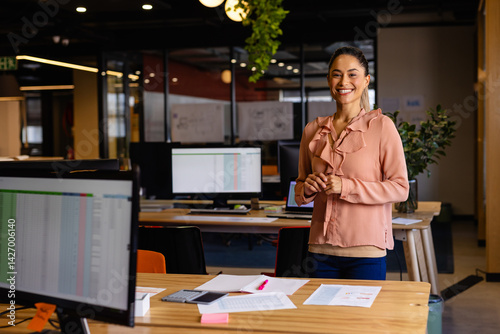 Obraz na płótnie Woman standing and smiling in modern office, with computer monitor and smartphone, copy space w salonie