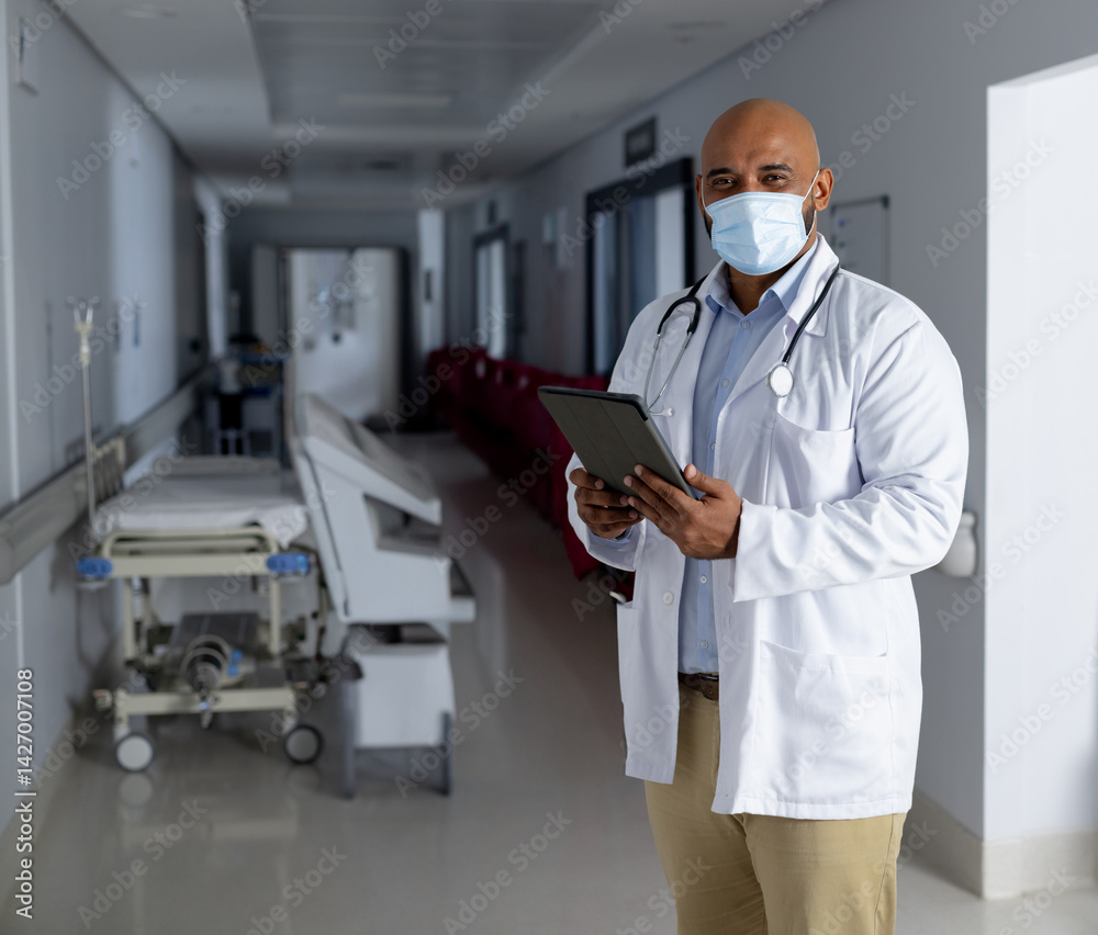 Male doctor reviewing tablet in hospital corridor, showing stethoscope and gurney, copy space