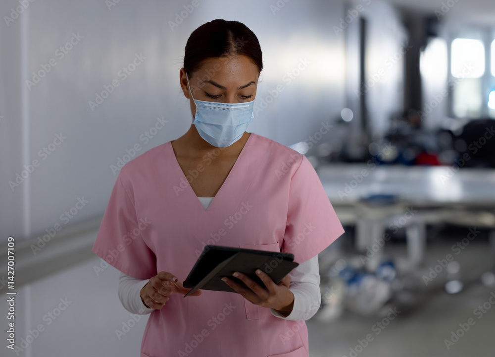 Female healthcare worker in pink scrubs studying tablet in hospital hallway wearing surgical mask