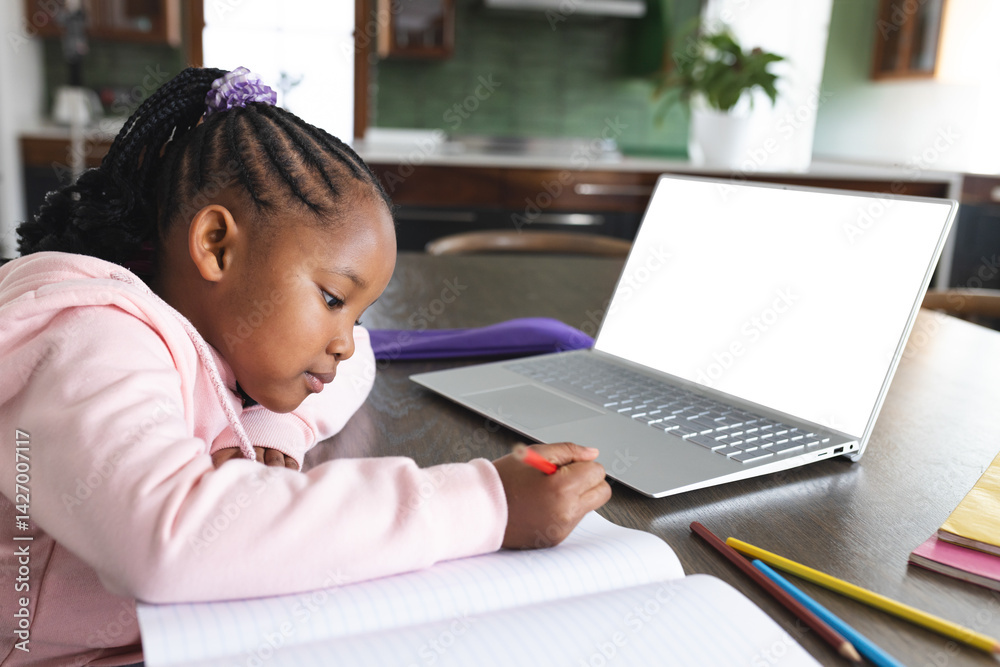 African American child writing at kitchen table with red pencil, open notebook, laptop, copy space
