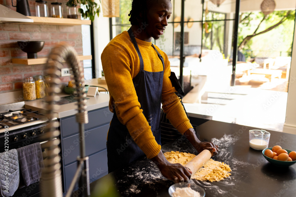 African American man rolling dough on floured kitchen island at modern home, with rolling pin