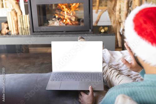 Obraz na płótnie Person wearing Santa hat sitting on sofa by fireplace, holding laptop with blanket draped over legs w salonie