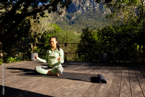 Obraz na płótnie Asian woman meditating on black yoga mat on wooden deck at mountain retreat, with rolled mat w salonie
