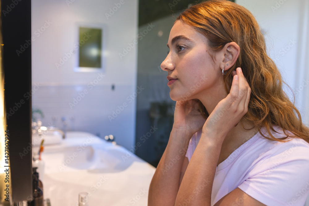 Woman adjusting earring and styling hair in home bathroom, with sink mirror and toiletry bottles
