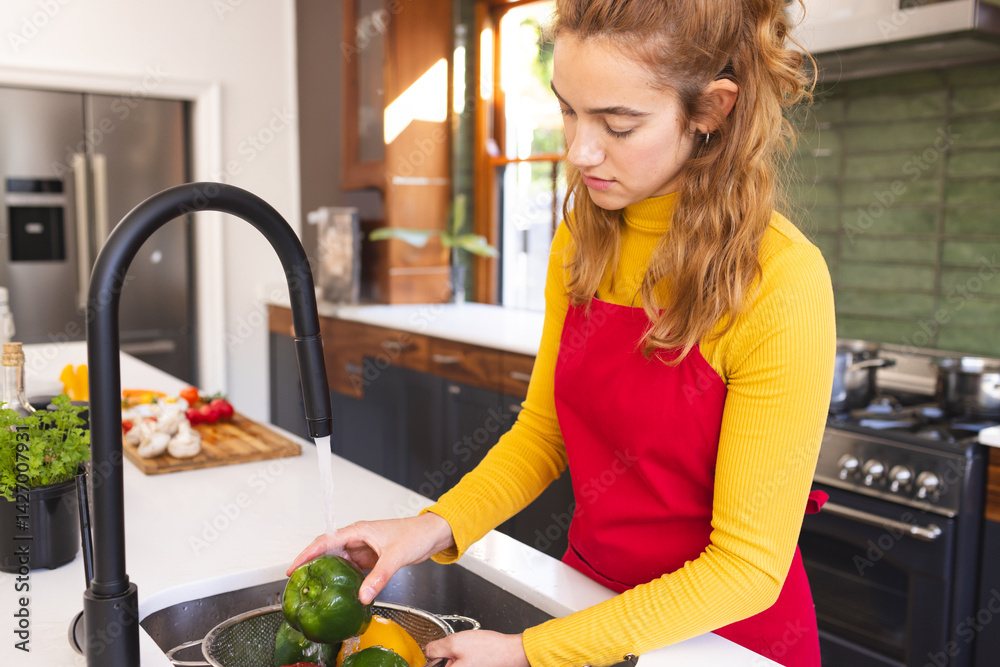 Young adult woman washing peppers in colander under black faucet at kitchen island, copy space