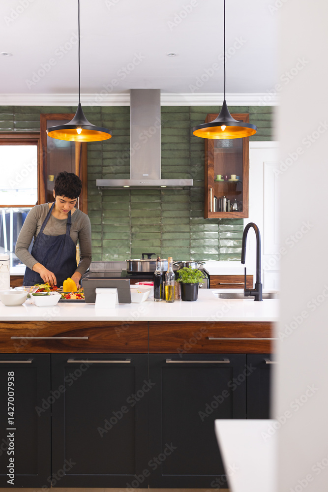 young adult female home cook chopping bell peppers on wooden cutting board in kitchen, using tablet