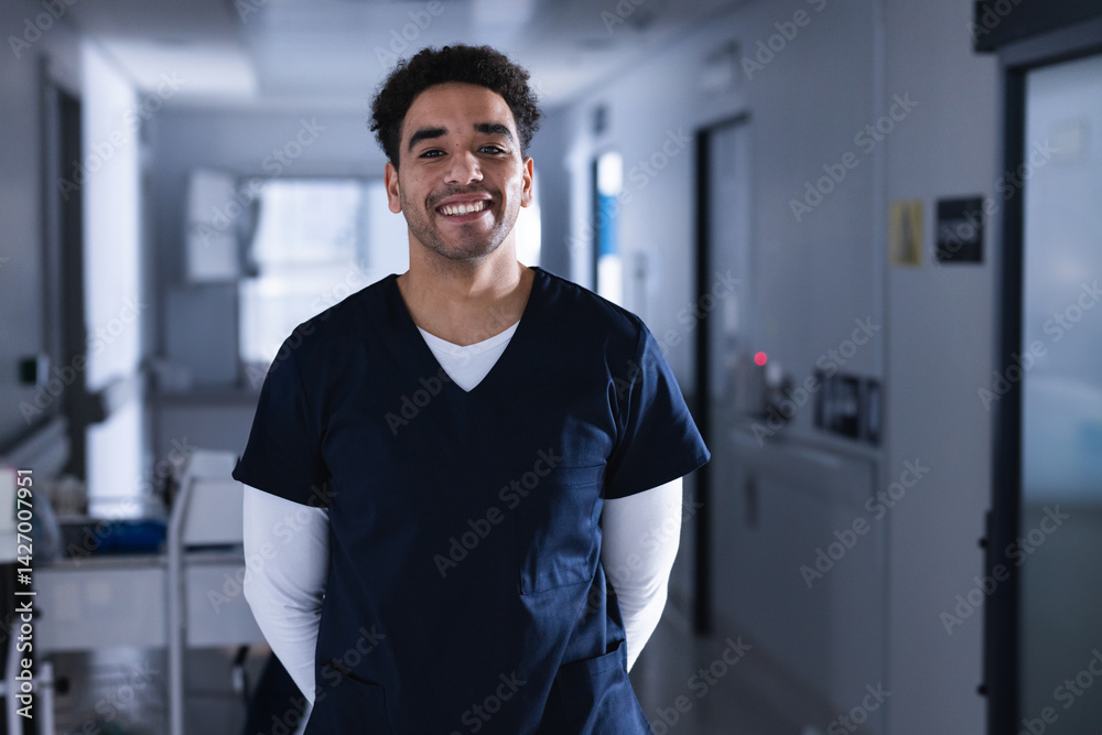 Young adult male healthcare worker standing in hospital corridor, smiling with medical cart behind