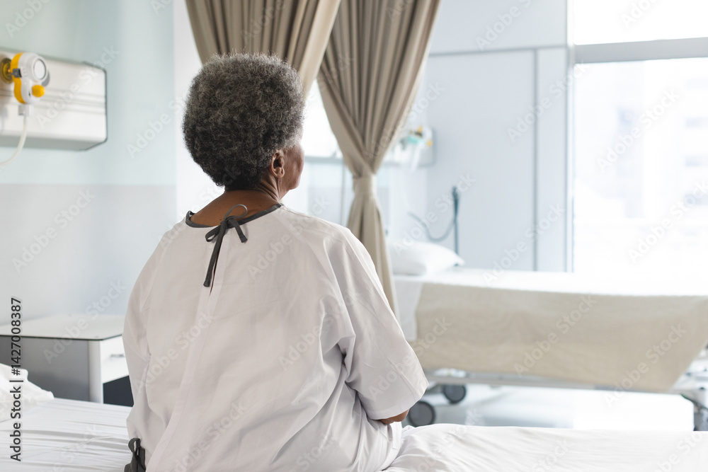 African American senior woman sitting on bed facing window in hospital room, with hospital gown