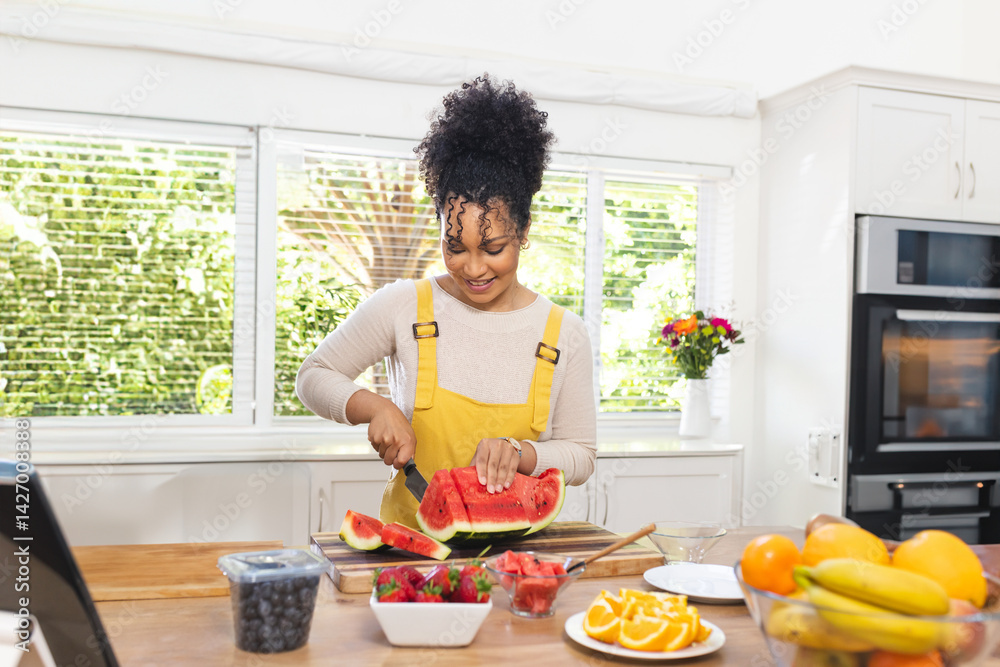 Woman slicing watermelon on cutting board on island in kitchen, with fruit bowl and tablet