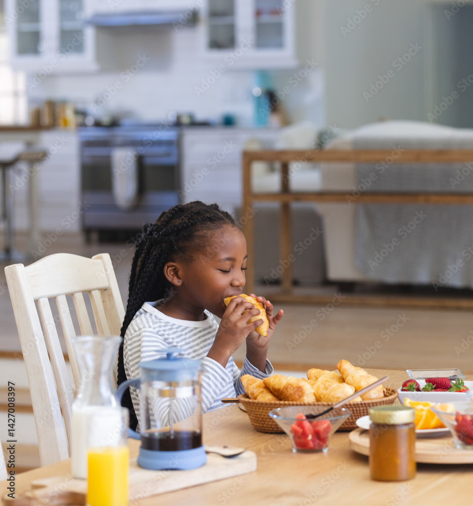 African American girl sitting at table in open kitchen eating croissant with bowl of strawberries