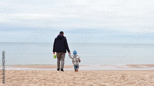 Father and son in autumn outfits walk hand in hand toward the sea on a sandy beach, capturing a calm family moment in soft coastal light.
