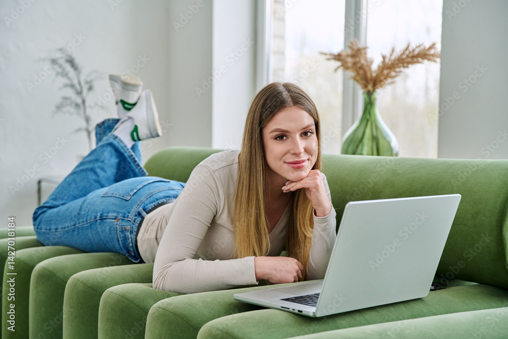 Portrait of young woman lying on sofa at home with laptop looking at camera