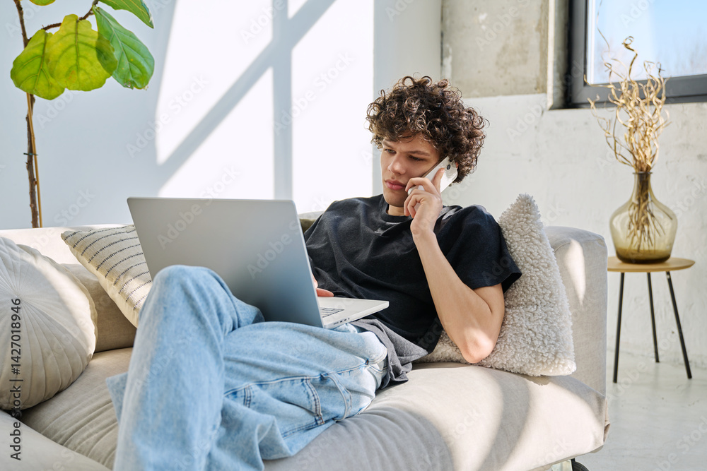 Handsome young man with laptop using smartphone sitting on sofa at home