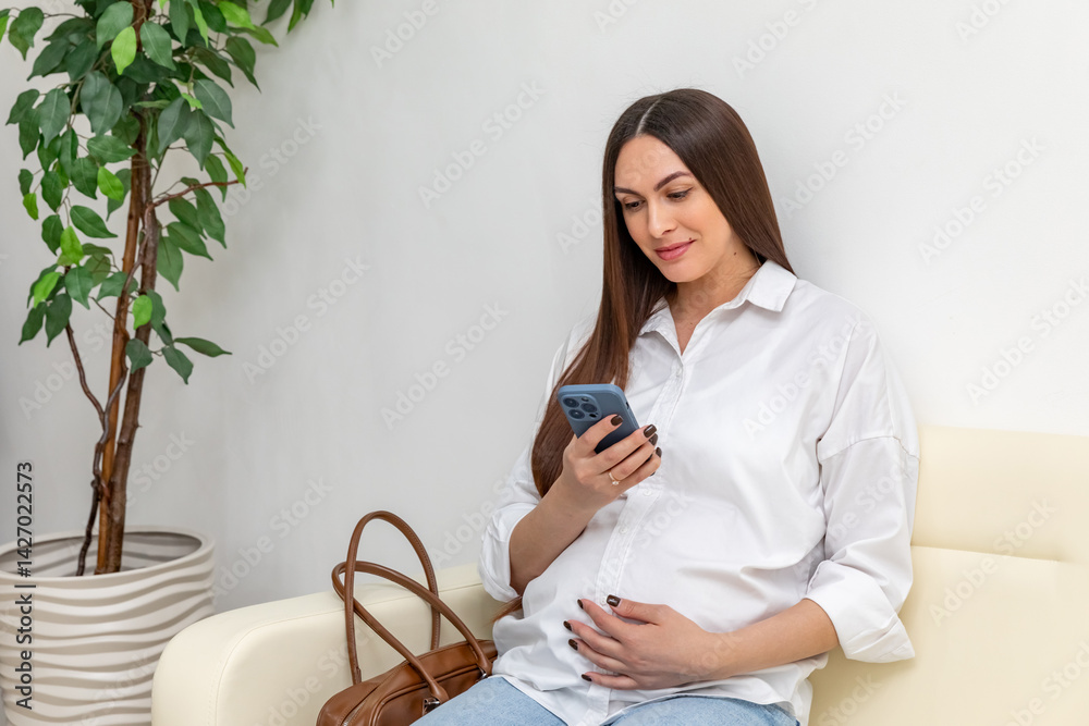 Pregnant woman using a smartphone and gently touching her belly while relaxing on couch in hospital. Happy young expectant mother. Family planning and IVF centers concept. High quality