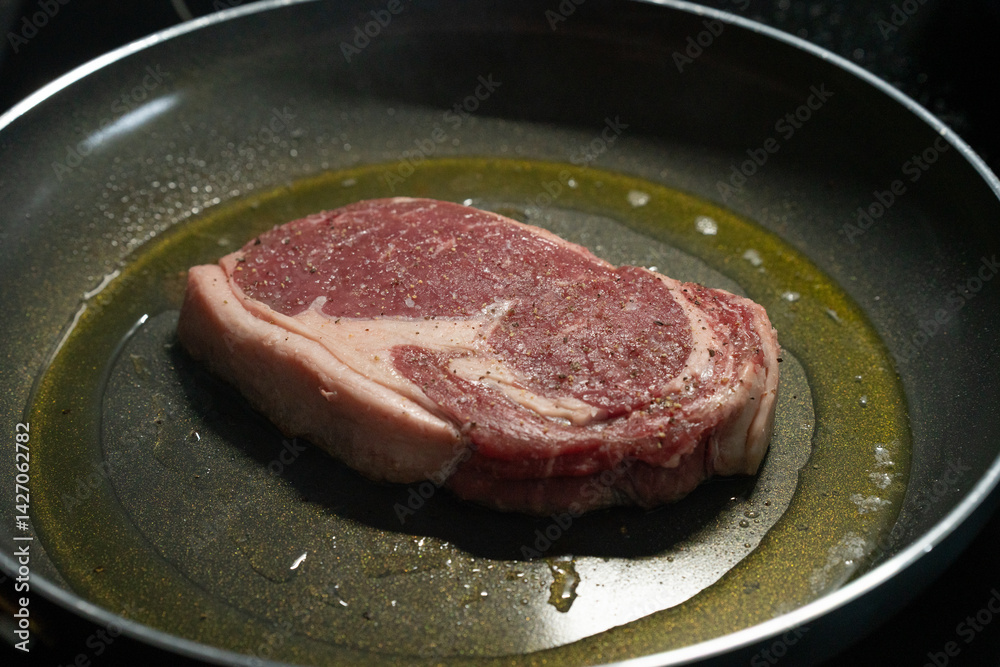 A ribeye steak sits in a hot skillet, surrounded by sizzling oil. The steak is seasoned and ready to be cooked, releasing flavorful aromas in the kitchen. A ribeye steak sits in a hot skillet, surrounded by sizzling oil. The steak is seasoned and ready to be cooked, releasing flavorful aromas in the kitchen.