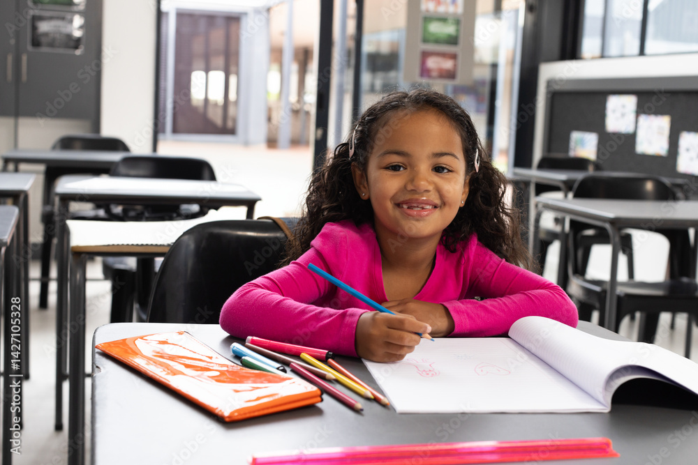 Girl drawing on white paper at modern classroom desk, using colored pencils and orange notebook