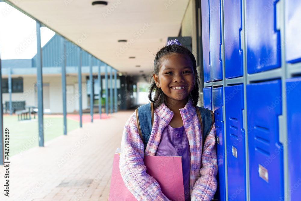 Multiracial girl leaning against blue lockers in school hall with binder and backpack, copy space