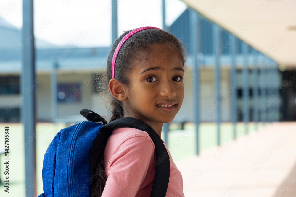 Girl child standing in covered school corridor, wearing pink headband and carrying blue backpack