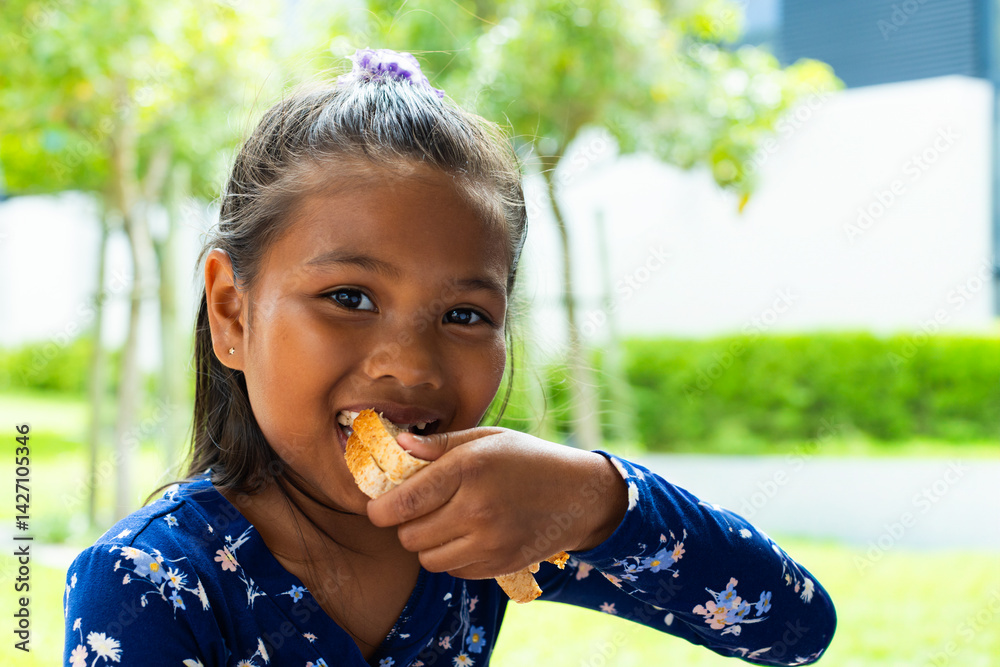 Young girl sitting outdoors in green backyard, holding torn bread piece and smiling