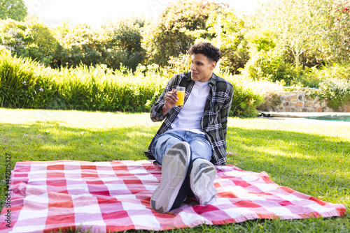 Obraz na płótnie Man sitting on red white checkered picnic blanket in backyard, holding glass of orange juice w salonie