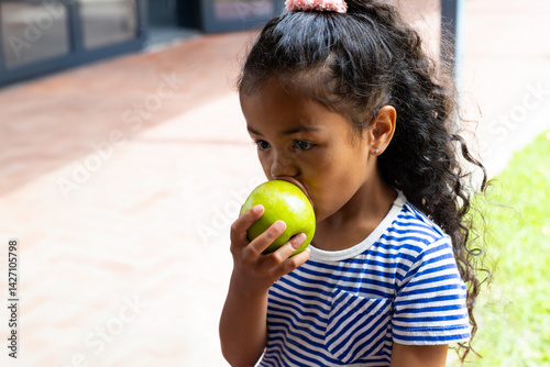 Obraz na płótnie Multiracial girl child biting bright green apple on brick patio at home, sunny snack moment w salonie